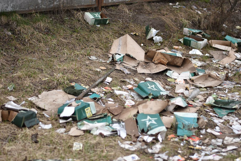 A patch of grassy ground near a fence is littered with a large amount of discarded cardboard boxes, many of which are crushed or torn, with some displaying visible brand logos and printed text. The debris also includes scattered paper waste, small plastic fragments, and broken packaging materials. The cardboard is primarily brown, with some pieces having a rough, corrugated texture, while others are smoother. The grass underneath is sparse and uneven, and the area appears to be an outdoor environment, possibly a backyard or side alley. In the background, a wooden fence is partially visible, with some soil and dried vegetation along its base. The scene suggests a site where waste has been hastily or haphazardly dumped, illustrating an untidy and unmanaged rubbish accumulation that could be addressed through sufficient rubbish removal procedures, such as those provided by House Clearance Maida Vale, to facilitate proper waste disposal or recycling.