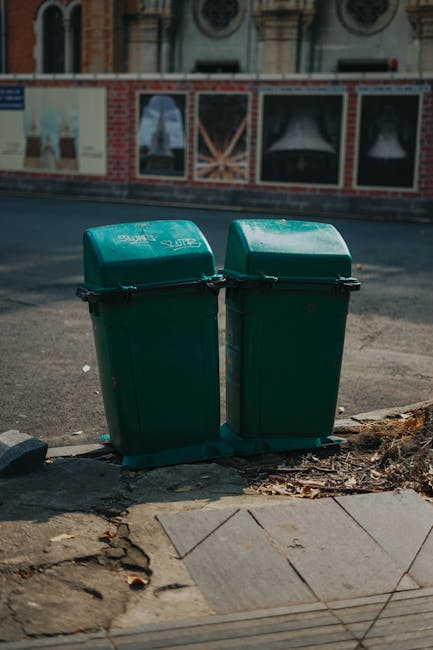 Two green wheeled rubbish bins with black handles and latches, positioned side by side on the pavement at the edge of a sidewalk. The bins are made of durable plastic with smooth, slightly reflective surfaces, and are labeled with white text indicating they are for waste collection. Behind the bins, there is a dark asphalt road with some small scattered debris at the curb. In the background, a brick wall with large, framed historic posters or advertisements, featuring images and text, is visible along with some architectural details of a street scene. The lighting is natural daylight, casting soft shadows on the ground, and the setting appears to be an urban residential area, typical of private waste collection or alternative rubbish disposal methods. The scene suggests an emphasis on independent waste management options, such as private rubbish removal services, aligning with the context of avoiding council-led rubbish collection fees, as discussed on the website houseclearancemaidavale.co.uk.