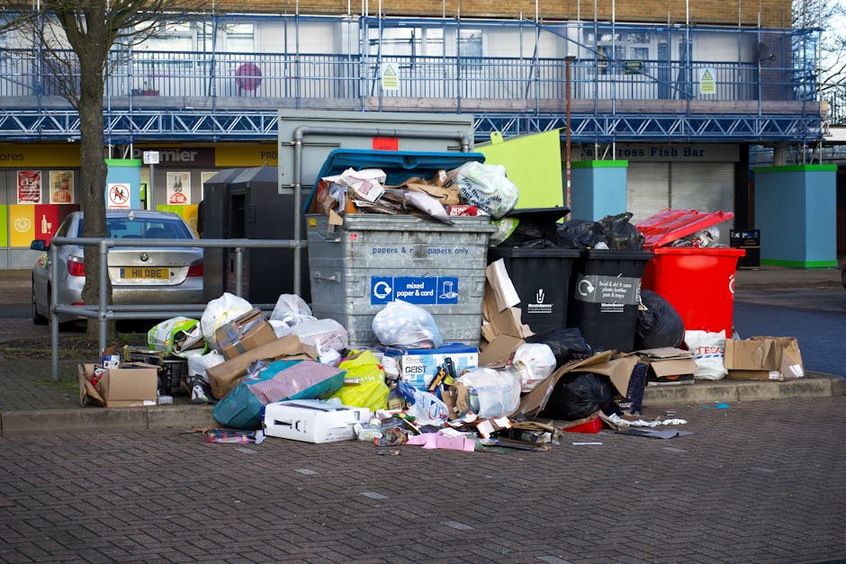 A collection of overflowing rubbish bins and discarded waste bags located on a paved area in an urban environment. The scene includes a grey mixed paper and cardboard recycling container with its lid open, revealing crumpled paper and packaging inside. Surrounding it are numerous black and black plastic rubbish bags, some tied and others torn open, spilling various waste items onto the ground. There are cardboard boxes, flattened and crumpled, along with miscellaneous packaging materials and paper clutter scattered around the bin area. To the left, a small silver car is parked adjacent to a metal railing, with a tree partially obscuring the vehicle. In the background, a building under construction or renovation is visible, wrapped with blue scaffolding and protective netting, with signage indicating commercial premises on the ground floor. The overall scene depicts a typical urban rubbish collection point awaiting private waste removal, with visible debris and waste materials in need of clearing by an expert rubbish removal service like House Clearance Maida Vale, consistent with independent waste handling practices in Maida Vale.