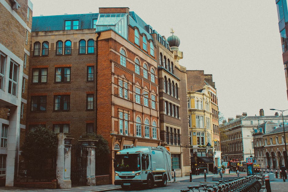 A narrow city street lined with multi-story buildings featuring a mix of brick and ornate architectural details, including arched windows and decorative stonework. The building on the left has a prominent corner with a small gate and iron fence, while the central building displays a reddish-brown brick facade with large, multi-pane windows. Behind, additional buildings with varied facades, including a cream-colored structure with intricate window frames, extend into the distance. In the foreground, a light blue waste collection truck with a white cab is parked on the street, adjacent to black bollards and a row of bicycles. The scene is illuminated by natural daylight, with overcast skies contributing to soft lighting. The environment suggests an urban setting where private waste collection services, such as those offered by House Clearance Maida Vale, might be employed as an alternative to municipal rubbish removal, particularly in densely built areas with limited parking or loading space. The overall scene captures a typical street scene oriented around property clearance and waste management activities within a city context.