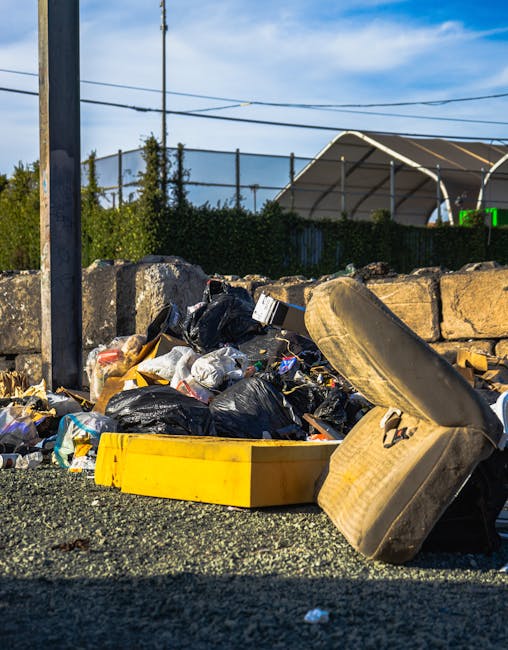 A collection of overflowing rubbish bins and discarded waste bags located on a paved area in an urban environment. The scene includes a grey mixed paper and cardboard recycling container with its lid open, revealing crumpled paper and packaging inside. Surrounding it are numerous black and black plastic rubbish bags, some tied and others torn open, spilling various waste items onto the ground. There are cardboard boxes, flattened and crumpled, along with miscellaneous packaging materials and paper clutter scattered around the bin area. To the left, a small silver car is parked adjacent to a metal railing, with a tree partially obscuring the vehicle. In the background, a building under construction or renovation is visible, wrapped with blue scaffolding and protective netting, with signage indicating commercial premises on the ground floor. The overall scene depicts a typical urban rubbish collection point awaiting private waste removal, with visible debris and waste materials in need of clearing by an expert rubbish removal service like House Clearance Maida Vale, consistent with independent waste handling practices in Maida Vale.