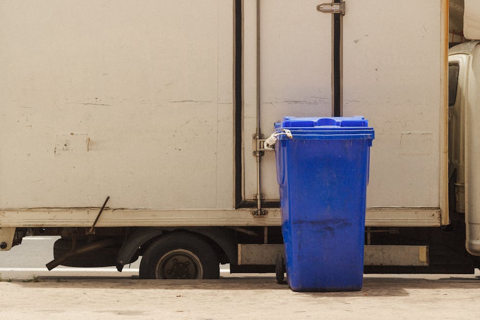 A blue wheeled rubbish bin made of plastic with a textured surface and a closed lid is positioned on a paved surface in front of a white delivery van or storage truck. The bin appears slightly dirty with visible black smudges and scuff marks on its surface. The van's side panel behind the bin is plain with some minor scratches and signs of wear, and it features a vertical metal handle or locking mechanism near the top. The lower section of the van includes a visible wheel arch and a small part of the wheel beneath the cargo area. The scene is illuminated by natural daylight, casting soft shadows and highlighting the textures of the bin and van. This setting suggests an environment where waste collection or rubbish removal services, like those offered by House Clearance Maida Vale, might occur, with the blue bin ready for lifting or disposal as part of private or alternative waste handling arrangements.