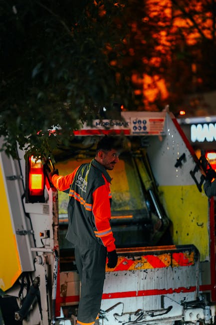 A waste management worker dressed in high-visibility orange and dark grey uniform with reflective stripes is seen standing by a large, yellow and white rubbish collection vehicle at dusk. The worker appears to be inspecting or preparing the vehicle, which is parked on a paved surface adjacent to a residential area. The back of the vehicle is open, revealing a compartment for waste, with some tools or equipment visible inside. In the background, the sky displays a deep orange hue, contrasting with the darkening environment, while a leafy tree partly obscures the upper part of the vehicle. The scene is illuminated by artificial lighting from the vehicle's signals, emphasizing the professional, on-site waste collection activity. The image naturally aligns with rubbish removal and private waste handling services offered by House Clearance Maida Vale, illustrating an example of independent waste collection process in an urban setting.