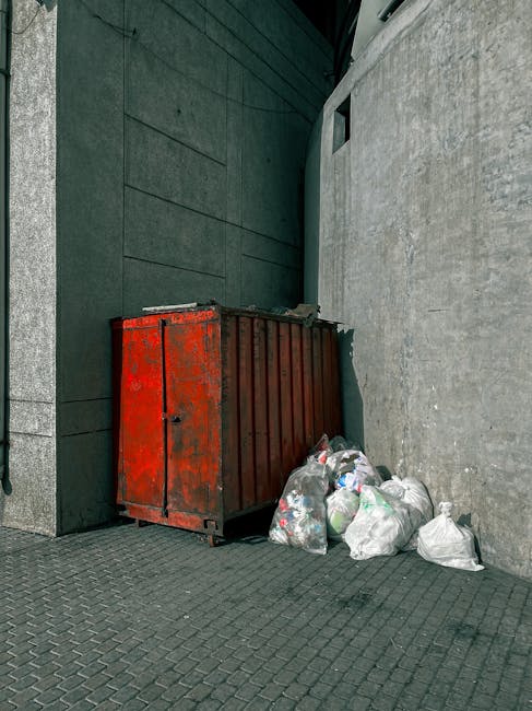 A large, weathered red metal skip positioned in a narrow outdoor alleyway corner beside a greenish-gray building wall with a smooth, dark surface and paneling. Adjacent to the skip are several partially filled white plastic bags containing mixed waste, including visible plastic bottles and packaging, resting on gray paving stones. The concrete wall on the right has a rough texture, while the sky is not visible, suggesting the scene is captured during daylight. The overall setting indicates a private waste disposal area, with the skip used for on-site rubbish collection, typical for independent disposal or clearance services such as those offered by House Clearance Maida Vale, highlighting practices distinct from local authority collection methods.