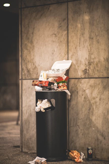 A tall, black cylindrical rubbish bin stands against a large, grey concrete wall in an urban environment. The lid of the bin is open, revealing a collection of mixed waste including paper, plastic, and food packaging, some of which has spilled onto the top of the bin and surrounding ground. Nearby, discarded trash such as crumpled paper, a drink can, and food wrappers are scattered on the pavement. The scene suggests casual littering or improper disposal, with various waste items resting on the ground and atop the bin. The lighting is natural but subdued, emphasizing the urban setting. The image visually relates to waste management and the importance of proper rubbish disposal, a subject covered by House Clearance Maida Vale’s services, including private and independent rubbish removal options outside of standard council collections.