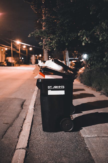 A black plastic rubbish bin with two wheels positioned on a sidewalk at night, illuminated by nearby streetlights. The bin is labeled 'St. John's' and is filled with various waste materials, including cardboard and plastic items, some of which are protruding from the open lid. The surrounding environment includes a quiet residential street with a row of trees and houses partially visible in the background, along with streetlights casting orange-yellow light and shadows on the pavement, indicating an evening setting. The scene suggests a private waste collection or rubbish removal process, potentially facilitated by services like House Clearance Maida Vale for non-standard or bulky waste disposal, especially in areas where on-site or independent rubbish collection is preferred or required.