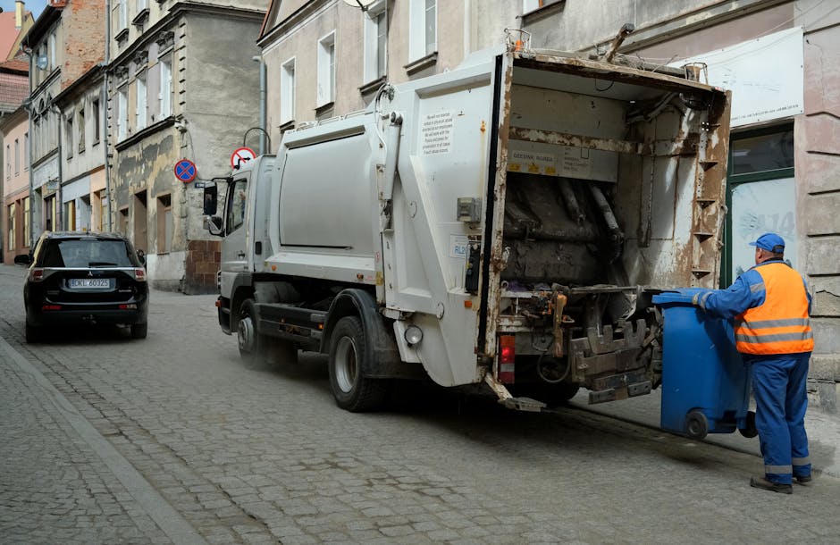 A large pile of mixed household waste and rubbish sits on a paved area in front of a multi-storey building, with the waste accumulated around and on top of several wheeled bins and trash containers. The waste includes crumpled cardboard boxes, plastic bags filled with rubbish, paper packaging, and discarded packaging materials, some spilling onto the surrounding pavement. The containers are made of plastic and metal, with some being black, red, or grey, and labelled for mixed paper and cardboard waste. In the background, there is a parked silver vehicle with a visible license plate, behind a metal safety barrier. The building behind the waste features blue scaffolding and a balcony, indicating ongoing maintenance or renovation work. The scene appears to be during daylight, with natural light illuminating the area, and the environment suggests an urban or commercial setting where independent rubbish collection may be arranged through services like House Clearance Maida Vale for non-standard or bulky waste disposal, supporting alternative waste handling methods outside local authority collections.