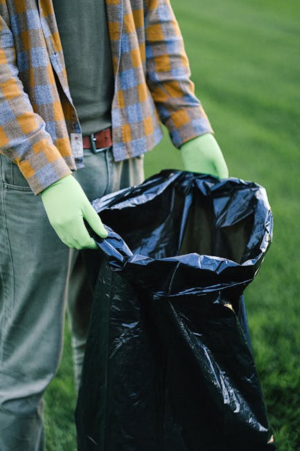 A person wearing a green long-sleeved shirt and a checked yellow and gray jacket is holding open a large black rubbish bag lined with a silver inner layer, prepared for waste disposal. They are standing outdoors on a grassy area, with a blurred background of green lawn or field. The individual is wearing light gray trousers with a brown belt and bright green gloves, indicating they are engaged in waste collection or rubbish removal work. The scene appears to involve private or on-site waste handling, typical of independent rubbish clearance activities. The black rubbish bag is partially filled, ready for rubbish disposal, and the natural outdoor setting suggests it could be part of a garden or yard clearance. This visual aligns with services provided by companies like House Clearance Maida Vale, which offer responsible waste removal solutions. The lighting is natural and even, highlighting textures of the gloves, the bag’s shiny surface, and the surrounding green environment, emphasizing an organized and professional approach to rubbish collection in an outdoor setting.