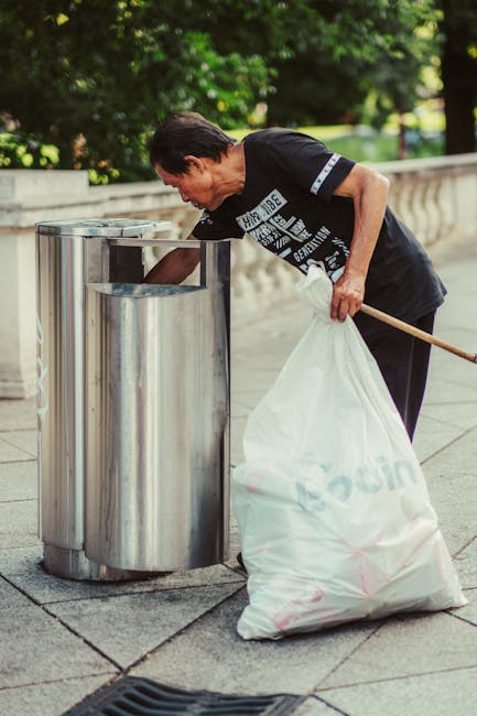 A middle-aged man with short dark hair and light skin is seen disposing of rubbish into a public waste receptacle on a paved outdoor area. He is wearing a black T-shirt with white graphic text on the front and a grey stripe on the sleeve, paired with dark trousers. The man leans forward slightly, holding a long-handled tool in his right hand and a large white plastic bag filled with rubbish in his left hand. The metal waste bin features two cylindrical openings for waste disposal, with a polished, reflective surface, positioned on a concrete surface with visible joints. In the background, dense green foliage and trees provide a natural setting, with a low stone balustrade running parallel along the edge of the paved area. The overall scene is well-lit with natural daylight, emphasizing the clean and orderly environment typical of urban public waste management, reflecting practices like on-site rubbish collection, which complement private disposal services provided by companies such as House Clearance Maida Vale involved in rubbish removal services.