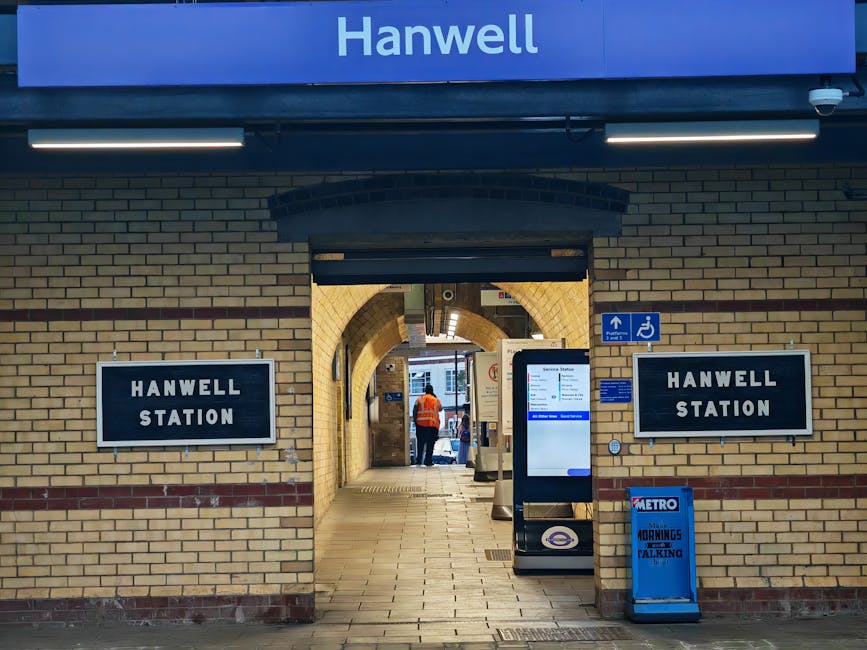 The image shows the entrance to Hanwell Station, constructed with light beige brickwork and a slightly arched brick archway over the passageway. On either side of the arch, there are rectangular black signs with white text that read 'Hanwell Station.' Above the arch, there is a blue sign with the word 'Hanwell' in white capital letters, illuminated by two linear light fixtures. To the right of the entrance, a small blue sign indicates accessible access for disabled passengers. In the foreground, a digital information screen mounted on a stand displays station details, and a blue Metro newspaper stand with a partially visible headline is placed to the right. Inside the station's entrance, a worker wearing an orange high-visibility vest is visible walking towards the platform, with additional interior signage and station features further in view. The scene suggests a typical urban train station setting with clean, well-maintained brickwork and modern signage, offering an environment where independent station access or alternative transportation options might be considered, which aligns with waste management services provided by House Clearance Maida Vale.
