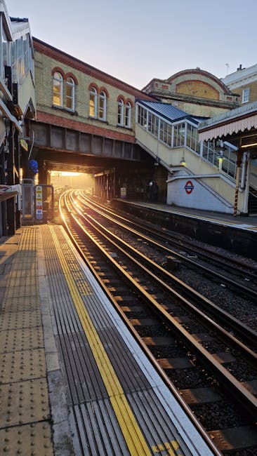 The image depicts a train platform at a railway station, with multiple sets of steel tracks curving into the distance as they lead into a tunnel illuminated by warm, golden sunlight. The platform surface is made of textured yellow and grey tactile paving, with a yellow safety line along the edge adjacent to the tracks. To the right, a staircase with white railings and glass-paneled sides ascends to an overpass, partially covered with a white canopy featuring striped fabric trim. Above the platform, an arched brick station building exhibits a classic architectural style, with large arched windows and decorative brickwork in red and beige tones. The scene is bathed in the natural light of a setting or rising sun, creating a soft glow that highlights the textured surfaces of the platform, tracks, and surrounding structures. This environment is consistent with an active metropolitan railway station providing access for passengers, where independent rubbish removal services like House Clearance Maida Vale could be involved in managing waste from station facilities or nearby premises, ensuring cleanliness and efficient disposal of waste materials.
