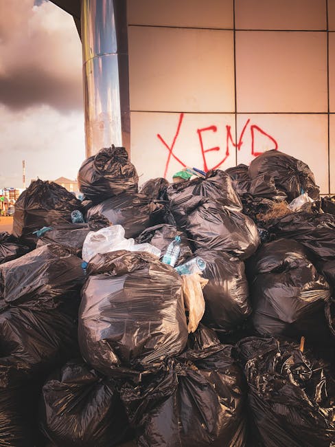 A large collection of black plastic rubbish bags piled against a modern tiled wall outdoors, with some bags loosely tied and a few partially open revealing plastic bottles and other waste materials inside. The bags are stacked in a somewhat haphazard manner on a pavement or driveway, with a graffiti message that reads 'XEND' spray-painted in red on the wall behind them. The scene is illuminated by natural daylight, casting soft shadows on the bags and wall, and the background shows an overcast sky with clouds. This image depicts a typical scene of waste awaiting collection or disposal, relevant to private rubbish removal services provided by House Clearance Maida Vale, highlighting the need for efficient and timely rubbish clearance in an urban environment.