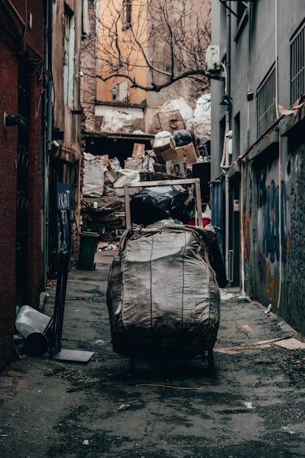 A large collection of black plastic rubbish bags piled against a modern tiled wall outdoors, with some bags loosely tied and a few partially open revealing plastic bottles and other waste materials inside. The bags are stacked in a somewhat haphazard manner on a pavement or driveway, with a graffiti message that reads 'XEND' spray-painted in red on the wall behind them. The scene is illuminated by natural daylight, casting soft shadows on the bags and wall, and the background shows an overcast sky with clouds. This image depicts a typical scene of waste awaiting collection or disposal, relevant to private rubbish removal services provided by House Clearance Maida Vale, highlighting the need for efficient and timely rubbish clearance in an urban environment.