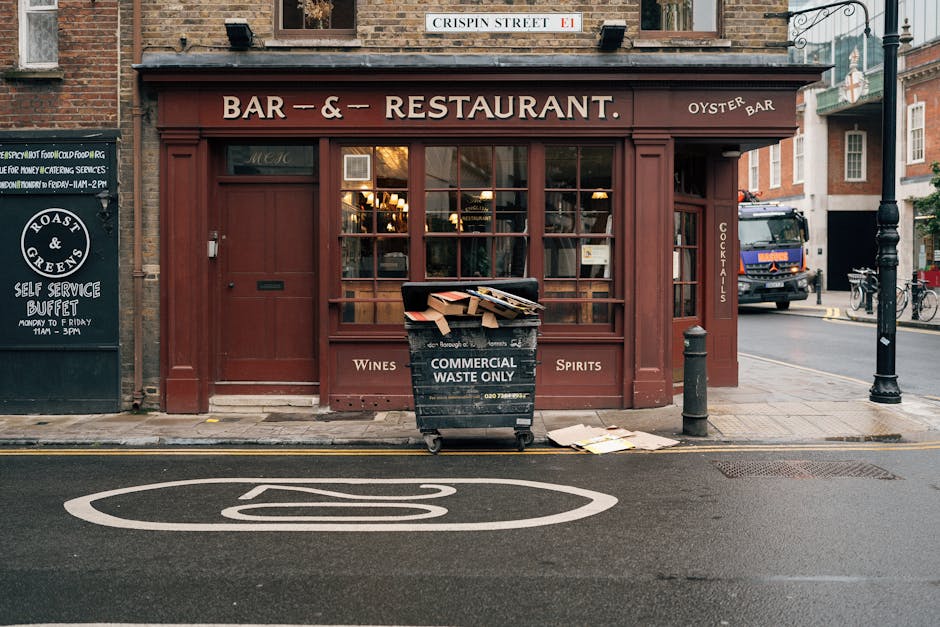 A street scene in front of a closed restaurant with a dark red wooden facade and large glass windows. The restaurant has signs indicating it sells wines and spirits, and features a vertical sign on the right side reading 'COCKTAILS.' In front of the establishment, a black wheeled bin marked 'COMMERCIAL WASTE ONLY' is overflowing with cardboard boxes, paper, and packaging materials, some of which are spilling onto the sidewalk. To the left, there is a small black chalkboard sign advertising a self-service buffet, and behind and to the right, a black bollard and street lamp are visible. A delivery truck is parked further down the street, alongside bicycles and residential buildings with brick exteriors. The scene is lit with natural daylight, and the road surface features painted markings, including a large, white, circular sign with a diagonal line, indicating no parking or stopping in this zone. The image illustrates an urban environment where private waste collection can be seen in relation to local commercial activity, aligning with independent rubbish removal services such as those offered by House Clearance Maida Vale. Considering the setting, the overflowing waste bin represents the need for proper rubbish disposal in compliance with waste rules for local tenants and businesses. The overall environment emphasizes street-level rubbish management, with waste left outside the premises awaiting collection.
